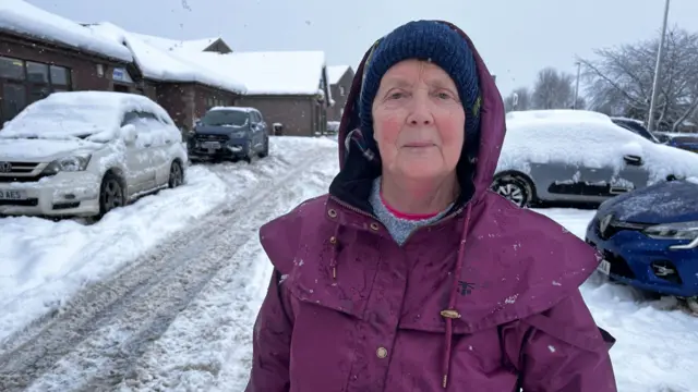 Eleanor Cardno wearing a purple jacket with the hood up covering part of her head. She is looking right at the camera and is also wearing a blue hat.