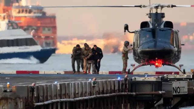 Maduro in the process of being transferred, there is a helicopter in the background and officials on either side of him