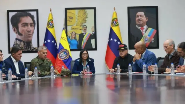 Delcy Rodriguez,  a woman with dark hair, sits at the end of a table with several Venezuelan flags around her. There are people sitting to either side, one in military fatigues, and others in suits. Pictures of past Venezuelan leaders are on the wall behind her
