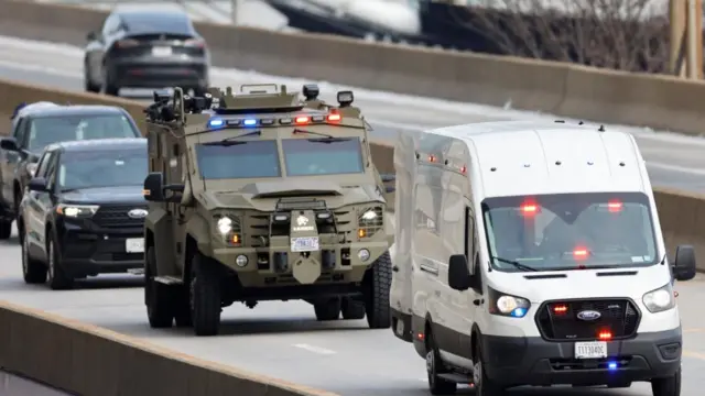 An armoured car with blue and red emergency lights pictured behind a white van with blue and red emergency lights