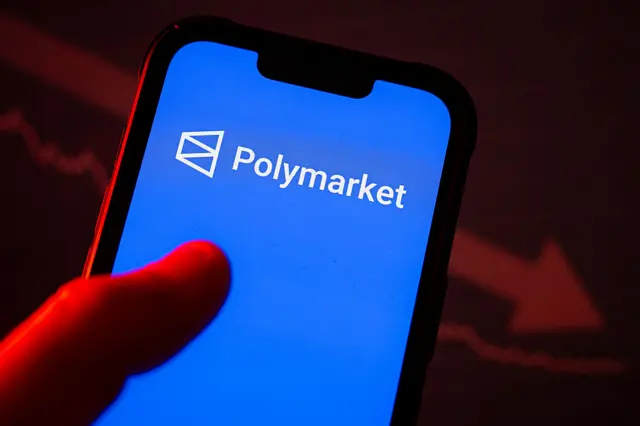 Thumb on top of a phone with a blue screen, the name Polymarket, and the company's logo - a trapezoid with a "v" in the middle on its side