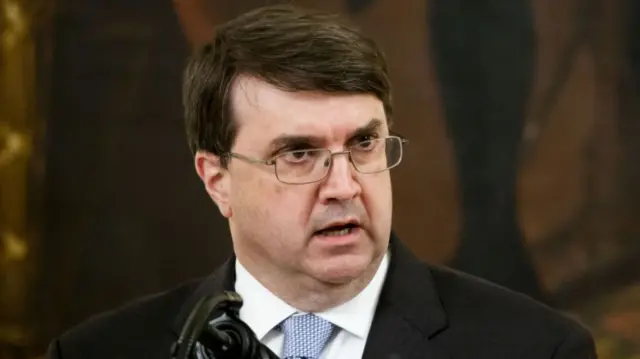 Robert Wilkie, a man with short brown hair wearing silver-rimmed glasses, a black suit jacket, a white shirt and a blue tie, speaks into a microphone.