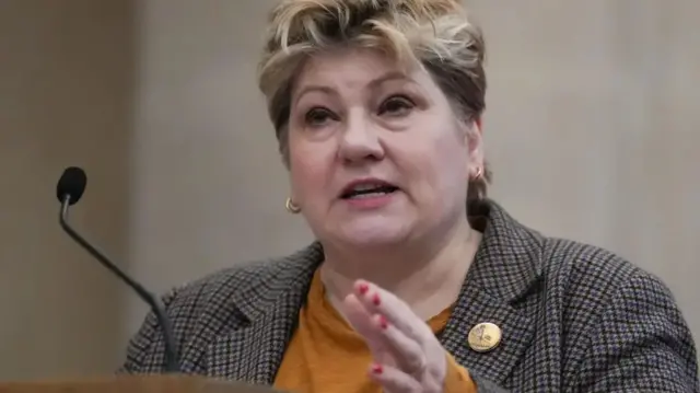 Dame Emily Thornberry wearing an orange t-shirt underneath a chequered suit, speaking at a podium with a microphone