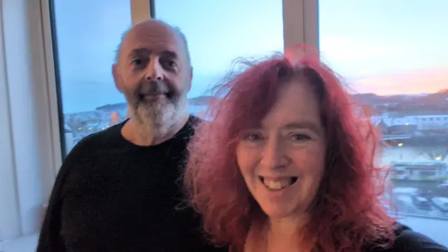 A middle-aged man with dark grey hair (L) and a smiling woman with red hair (R) take a selfie standing in front of a window. Behind him, a river view with the street and buildings covered in snow