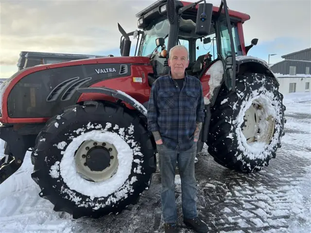 Duncan Wright wearing a dark blue flannel shirt, dark jeans and dark boots looking straight at the camera. He has white hair. His hands are in his pockets. He is stadning in front of a red tractor which is partially covered with snow on a snowy patch of ground.