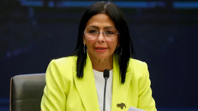 Delcy Rodriguez, a woman with long dark hair and gold hoop earrings, is wearing a bright yellow suit jacket with a pin of Venezuela on her lapel