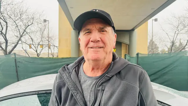 An elderly man in a dark zip-up jumper with a hoodie poses in front of a car under what appears to be a raised walkway