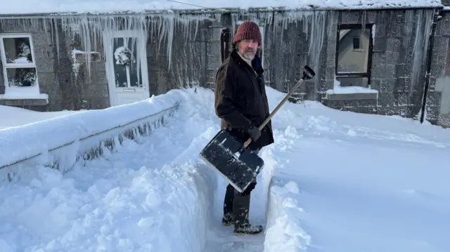 A man stands in path he has cleared in the snow. It is about thigh deep and he is wrapped up and holding a shovel. A house behind him has long icicles hanging off it