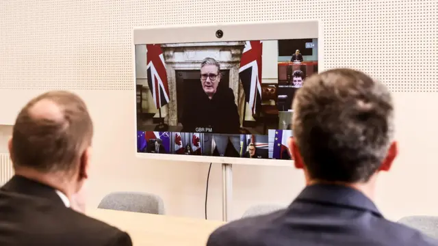 UK Prime Minister Keir Starmer speaks via videoconference at the Coalition of the Willing in December 2025. He's on a television screen surrounded by other people in the video call. In the foreground are two European leaders listening in.