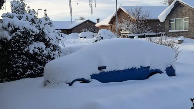 deep snow over a car on a driveway with houses in the background