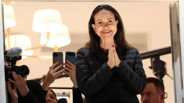 Machado standing on a balcony, wearing all black, smiling and with her hands in a prayer position.