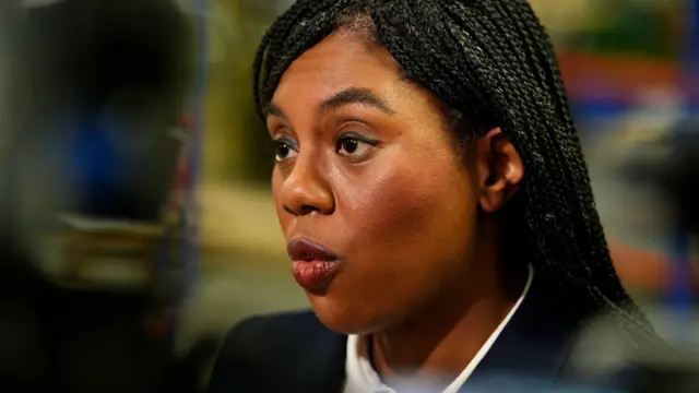Kemi Badenoch up close and looking to the left of the image. She is wearing a white collared shirt with a black blazer and her hair is down. The background is blurry.