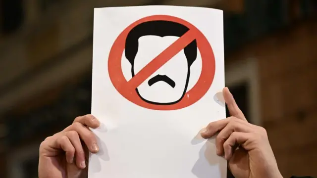 a pair of hands hold up a white sign with a cartoon outline of Maduro's face in a red circle and 'strike through' line