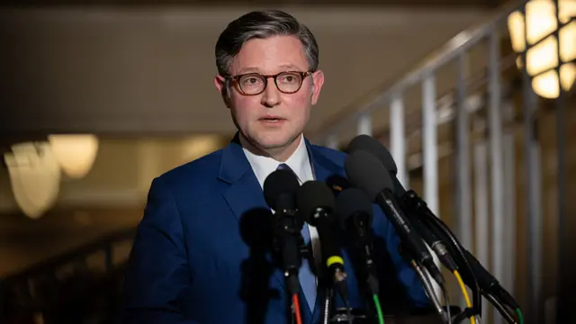 Mike Johnson speaking in front of microphones. He's wearing a suit and tie and glasses.