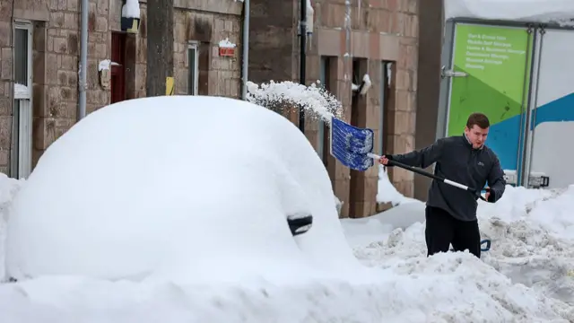 A man shovels snow in the street, next to a car covered in snow