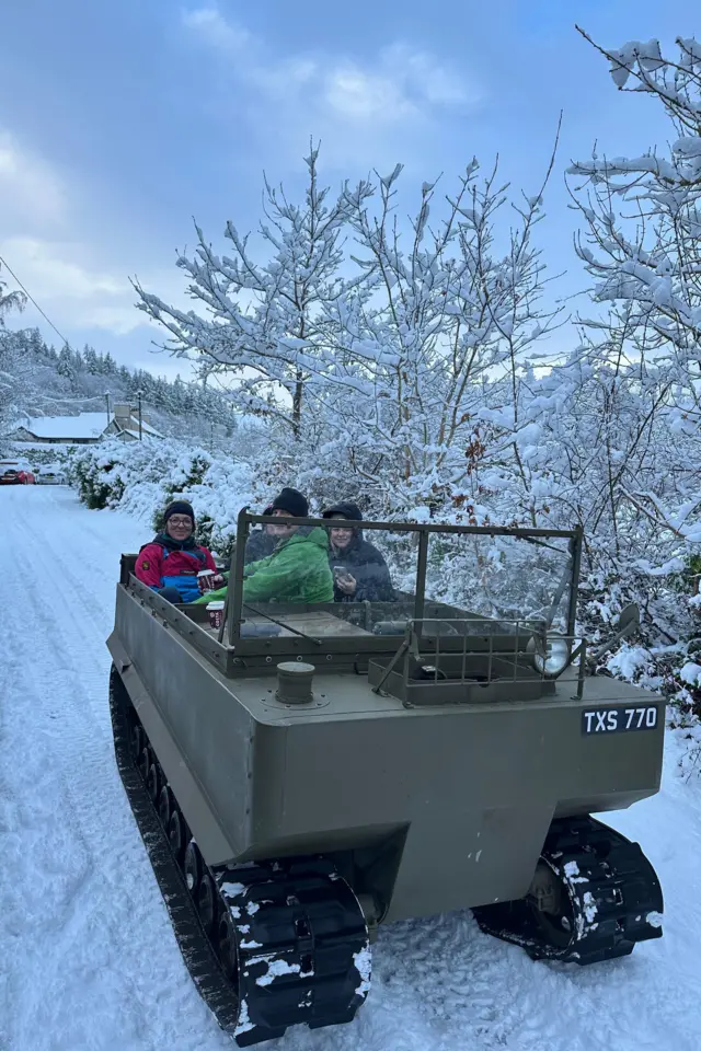 A world war 2 vehicle, with tank tracks, drives several people through the snow