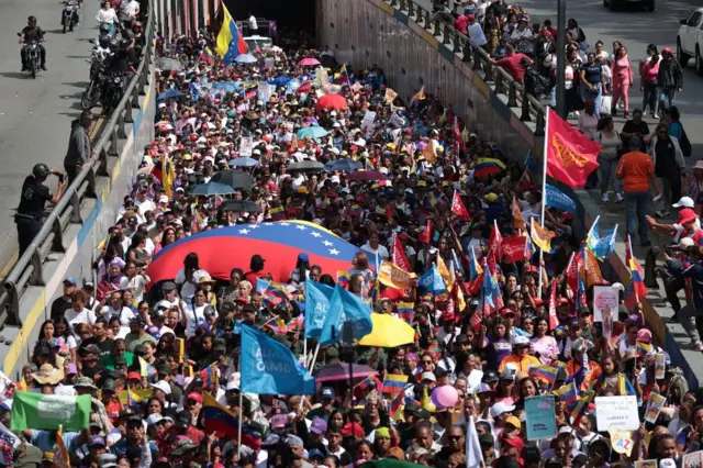 A large crowd waving Venezuela and other flags emerge from under an overpass bridge