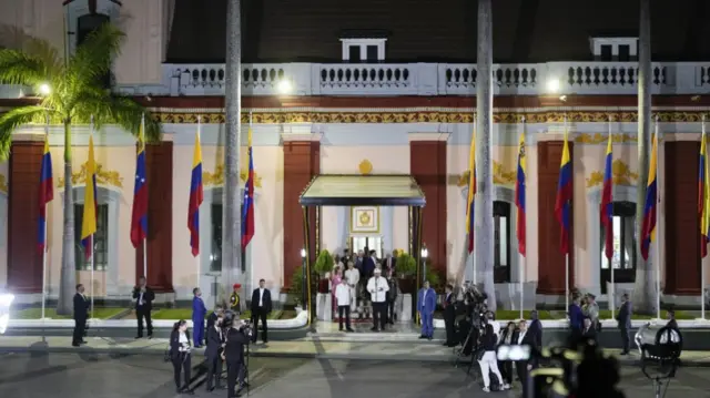 A press conference outside a regal building with lots of Venezuelan flags on the ground parallel to the building. A few reporters are stood on the pavement with cameras as a group of people stand at the door, giving the press conference.