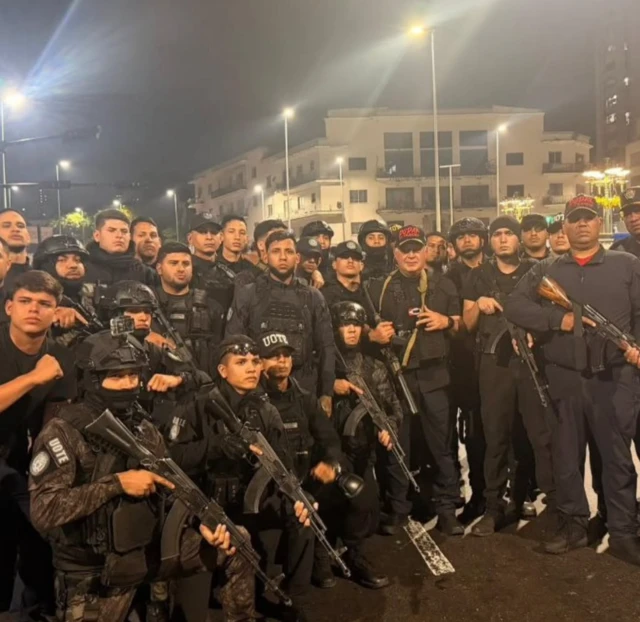 Lots of men with guns stand on the streets at night time posing for a photo