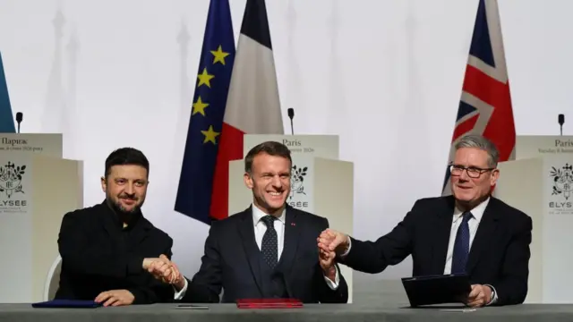 Ukrainian president Volodymyr Zelensky, French President Emmanuel Macron and UK Prime Minister Keir Starmer all shaking hands sat at a table as they sign the declaration