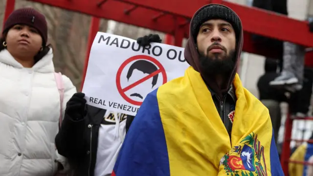 A protestor in New York City wrapped in the Venezuelan flag. Another holds a sign saying 'Maduro out', with a black and white outline of his face surrounded by a red stop symbol.