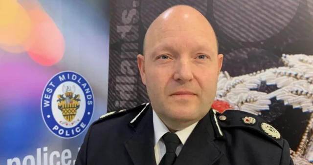 A man in a formal uniform looks at the camera, to the side of him is a sign saying West Midlands Police