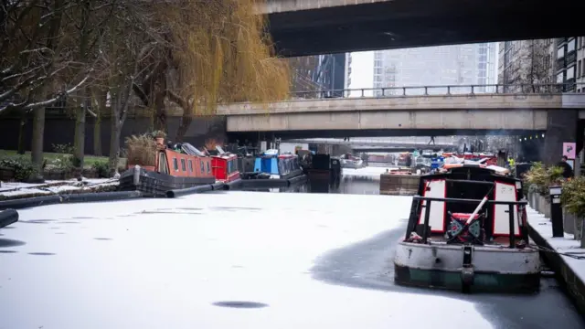 Several canal boats sit in frozen water, there are two bridges overhead
