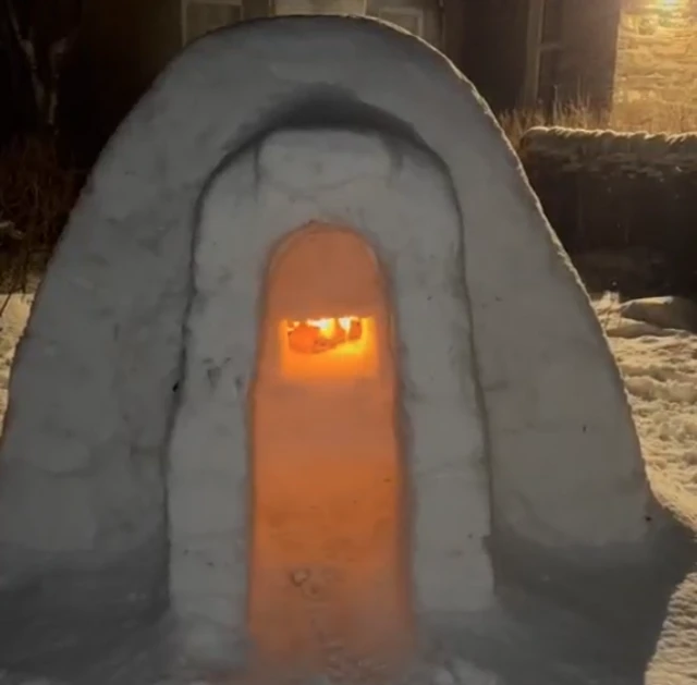 An igloo with a fireplace which glows amber. It has been built in a garden and it is dark outside