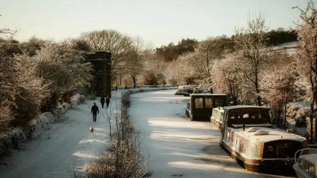 Boats covered in snow in a frosty canal, while four people and a dog walk along a nearby path covered in snow
