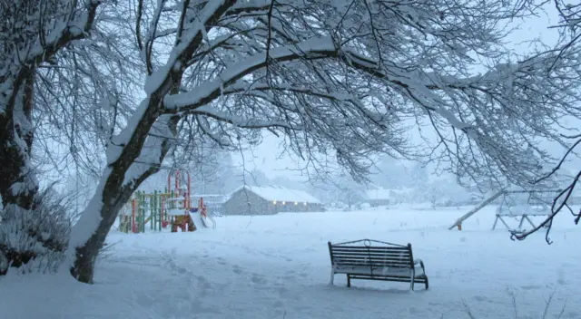 A bench in a snow covered field with a small building in the distance
