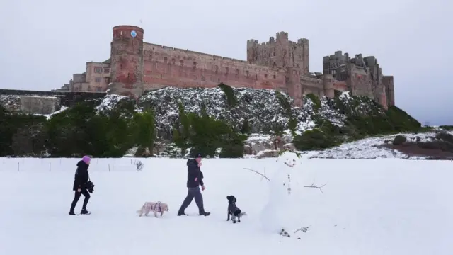Bamburgh Castle surrounded by snow in Bamburgh, Northumberland, there are two adults with two dogs and a snowman in front of the castle