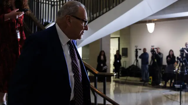 Schumer in jacket and tie and glasses on circular stairs with tv cameras in background