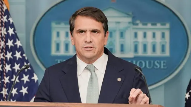 Deputy Attorney General Todd Blanche speaking to media from a lectern while dressed in a navy suit with a sage and white tie, with the White House sign visible behind him and the American flag to his left.