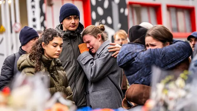 Young people cry and comfort each other in front of flowers at the scene.