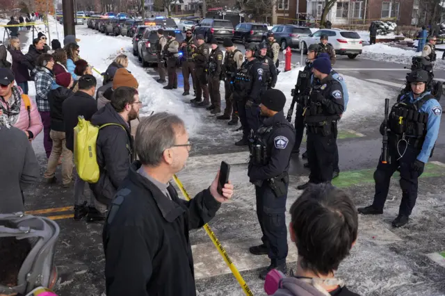 A crowd of people stand on one half of a yellow police cordon, while Minneapolis Police officers stand on the other