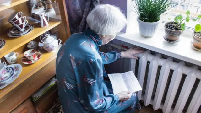 An elderly woman touches a radiator and holds a piece of papers
