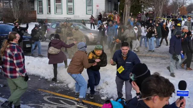 A crowd of reporters and the public stand in the snow and ice at the scene of the shooting