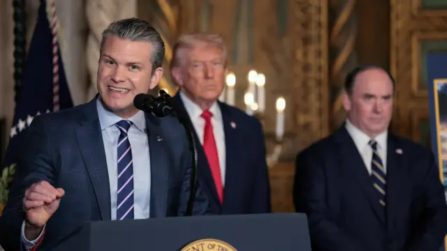 Pete Hegseth speaking at a microphone wearing a blue suit with Donald Trump behind him