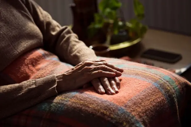An elderly woman with her hands clasped in her lap on top of a checked blanket. The image is a close up of her torso and hands and she is wearing a brown jumper and sitting next to a table with a phone on it and a plant