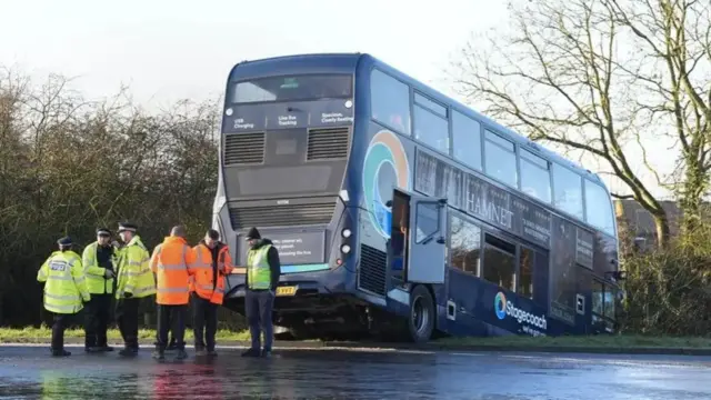 A blue Stagecoach bus has its front wheels in a ditch and its back door hanging open. Six emergency responders are speaking outside wearing yellow and orange high-vis vests.