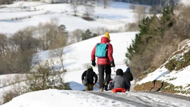 An adult in a red puffer jacket and blue backpack pulls a child on a red sled down a snowy hill.
