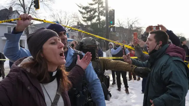 A woman, who is wearing a purple coat and hat, stands among a crowd beside yellow police tape