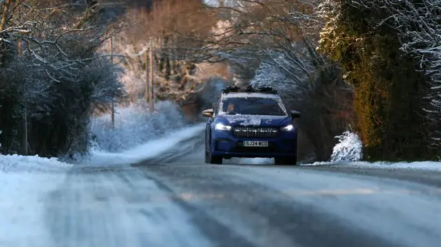 A motorist drives along a snow covered road. Snow can be seen on the front of the blue car
