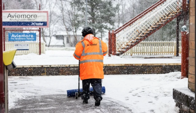 A ScotRail worker in an orange high-vis jacket, sweeping snow from the platform of a train station.