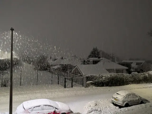 A street in Aberdeen with two cars, a few houses and a fenced off pitch covered in snow. A streetlamp is shining so we can see the snow falling heavily