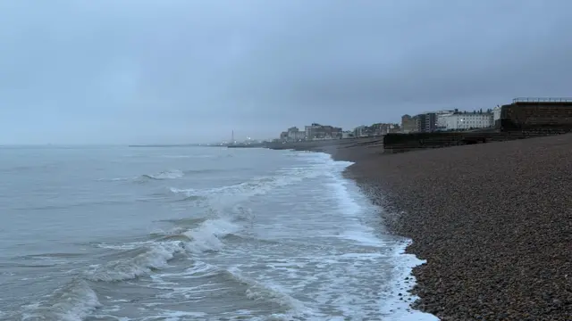 A coastline is pictured with small waves lapping at pebbles on the beach on an overcast morning