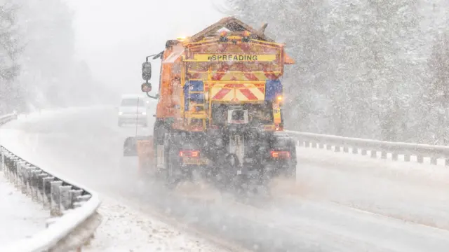 A yellow gritting truck travels along a main road in heavy snow. Oncoming traffic is visible through the murky, snowy sky.