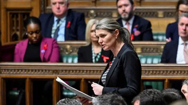 Jayne Kirkham MP is stood in parliament. She is wearing a black blazer and talking to other members.