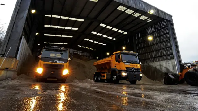 Two large yellow vans for gritting in a depot.