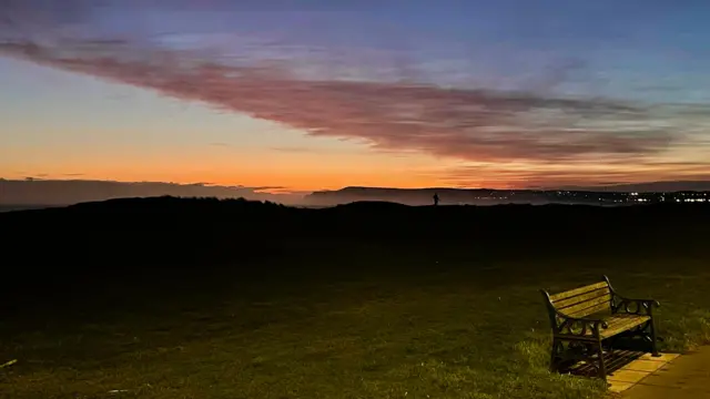 A sunrise is pictured with orange clouds and a park bench in the foreground on grass